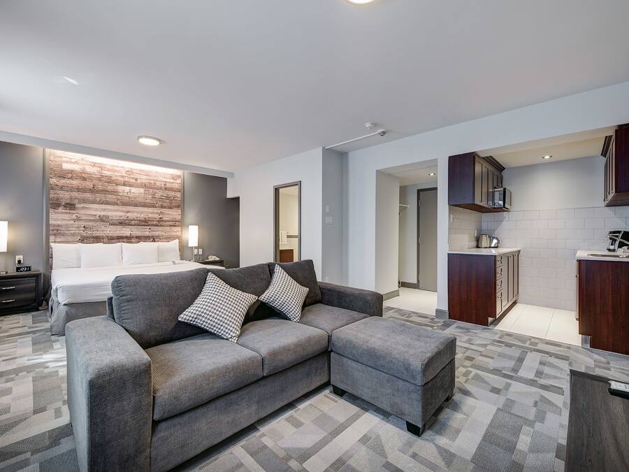 Une chambre d’hôtel moderne avec un canapé d’angle gris, un lit avec du linge de maison blanc, un mur d’accent en bois, une kitchenette avec des meubles en bois foncé et un tapis à motifs.