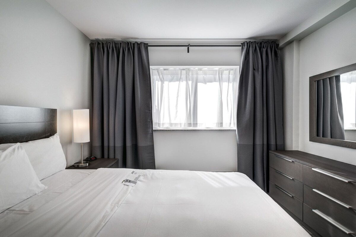 A hotel room with a bed, a bedside table with a lamp, a dresser, and a window with curtains.
