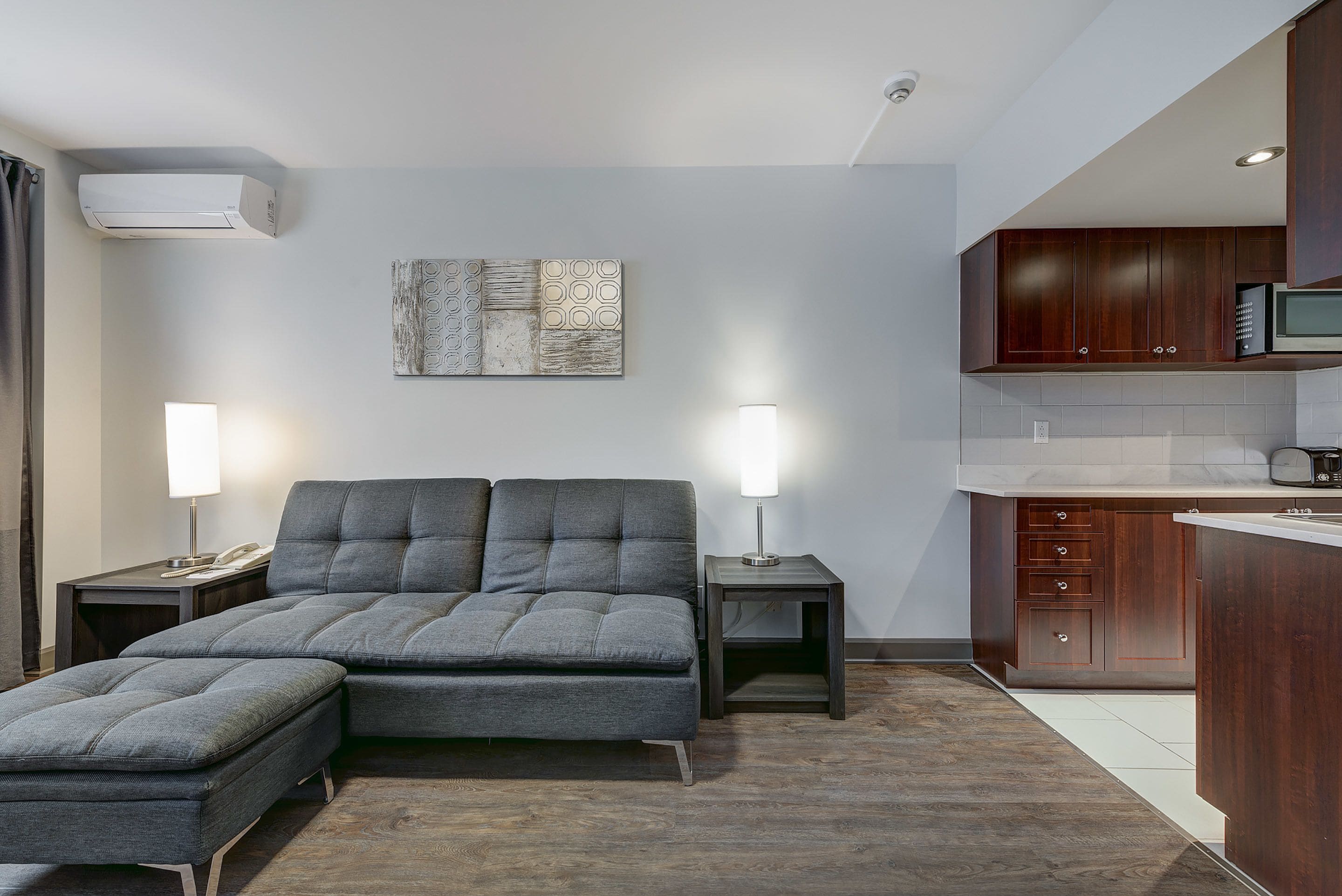 Una sala de estar moderna con un sofá y un reposapiés, una zona de cocina con armarios de madera y un aire acondicionado instalado en la pared.