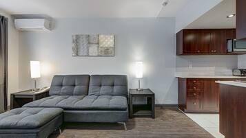 Una sala de estar moderna con un sofá y un reposapiés, una zona de cocina con armarios de madera y un aire acondicionado instalado en la pared.