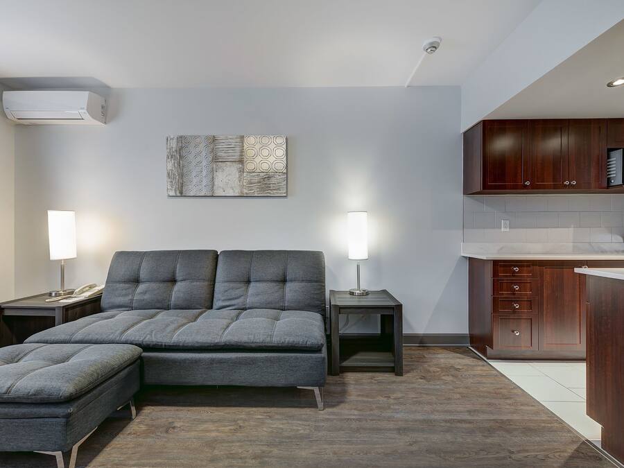Un salon moderne avec un canapé et un pouf, un coin cuisine doté de meubles en bois et un climatiseur fixé au mur.