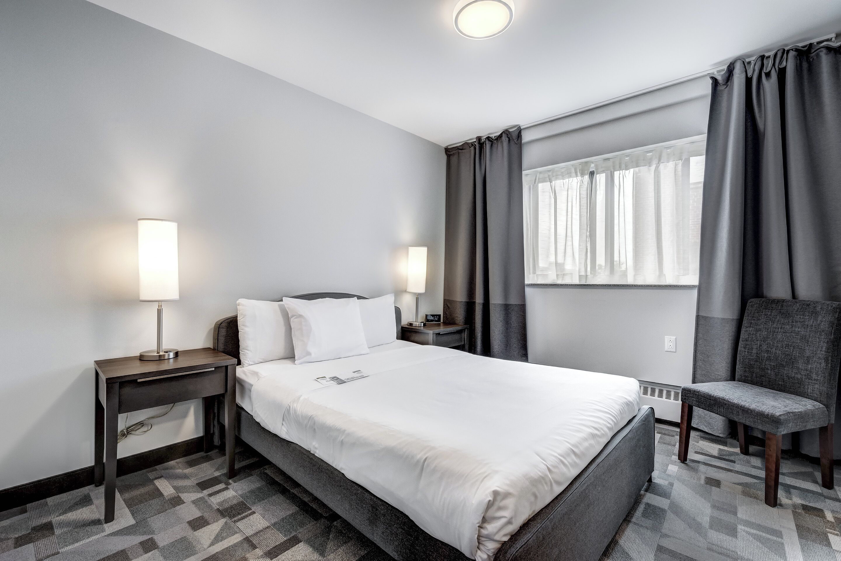 Une chambre d’hôtel comprenant un lit, deux tables de chevet avec des lampes, une chaise, une fenêtre avec des rideaux et un radiateur.