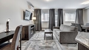 A modern living room with a sofa, coffee table, and a TV mounted on the wall. There is a desk with a lamp and a chair. The room has large windows with curtains. - Hotel Terrasse Royale (Montreal)