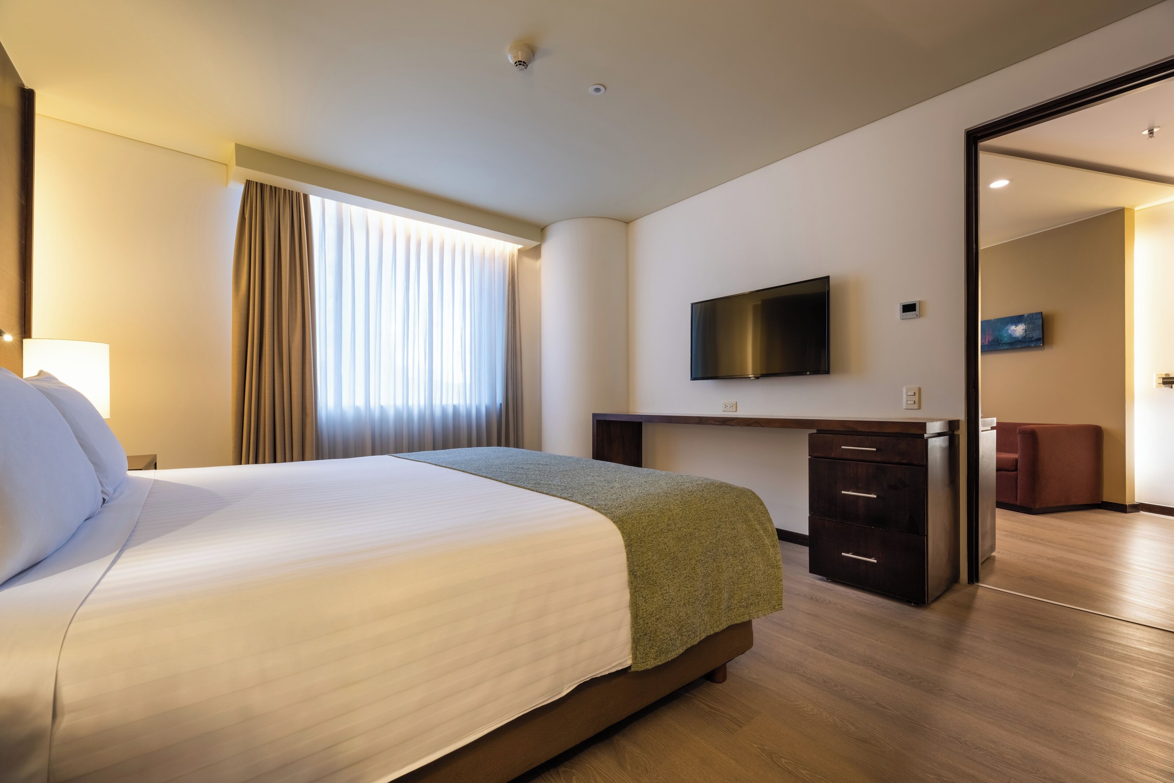 Habitación de hotel con cama, escritorio, televisor y puerta corredera que da a un área de descanso.