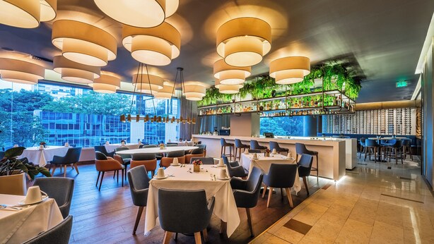 Un restaurante con varias mesas, lámparas colgantes, una zona de bar y vistas al exterior.