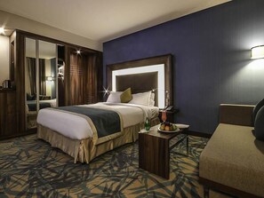 Quarto de hotel com uma cama grande, mesa de cabeceira com abajur, uma cadeira, espelho e guarda-roupa.