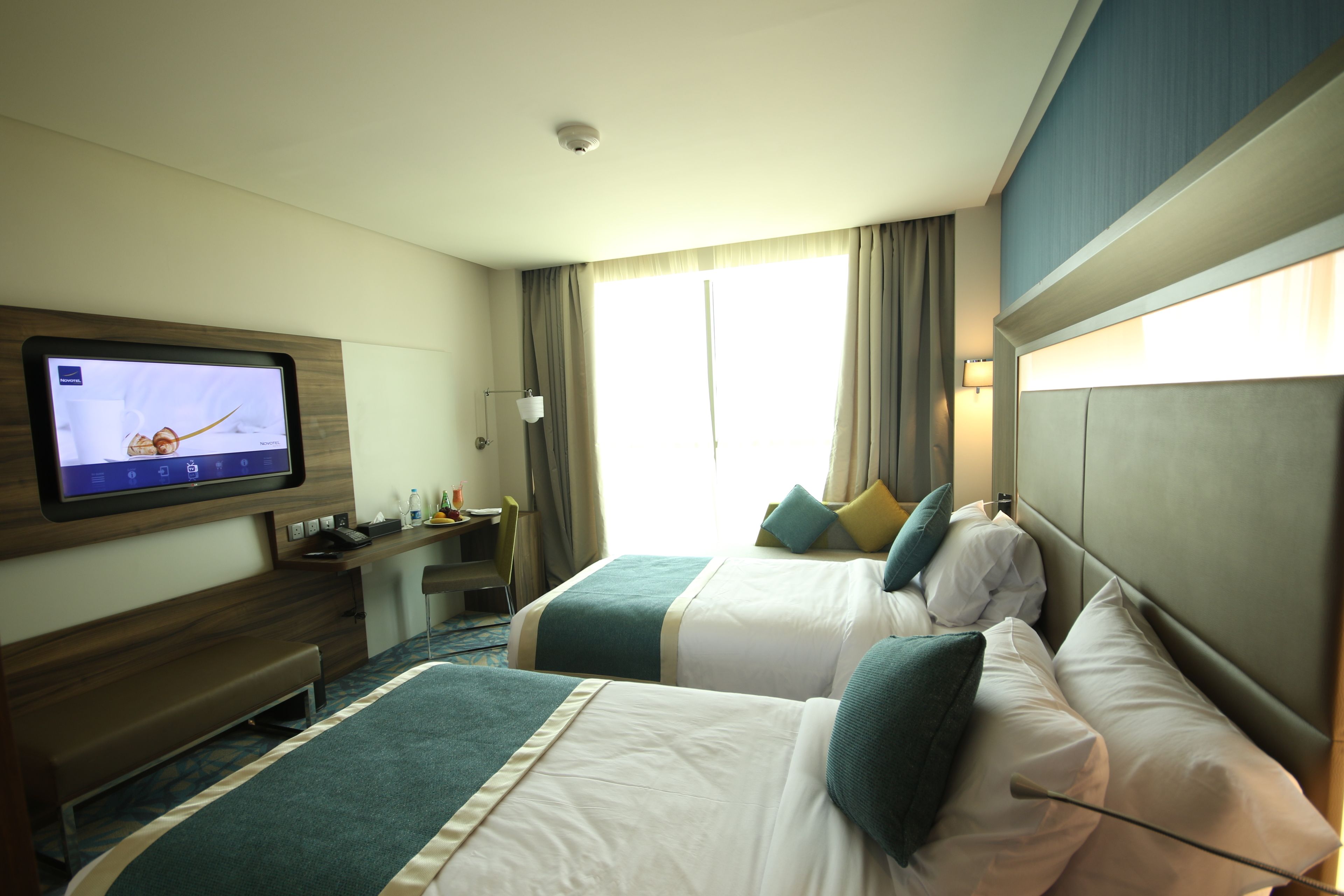 Een hotelkamer met twee bedden, een bureau met een computer, een televisie en een raam met gordijnen.