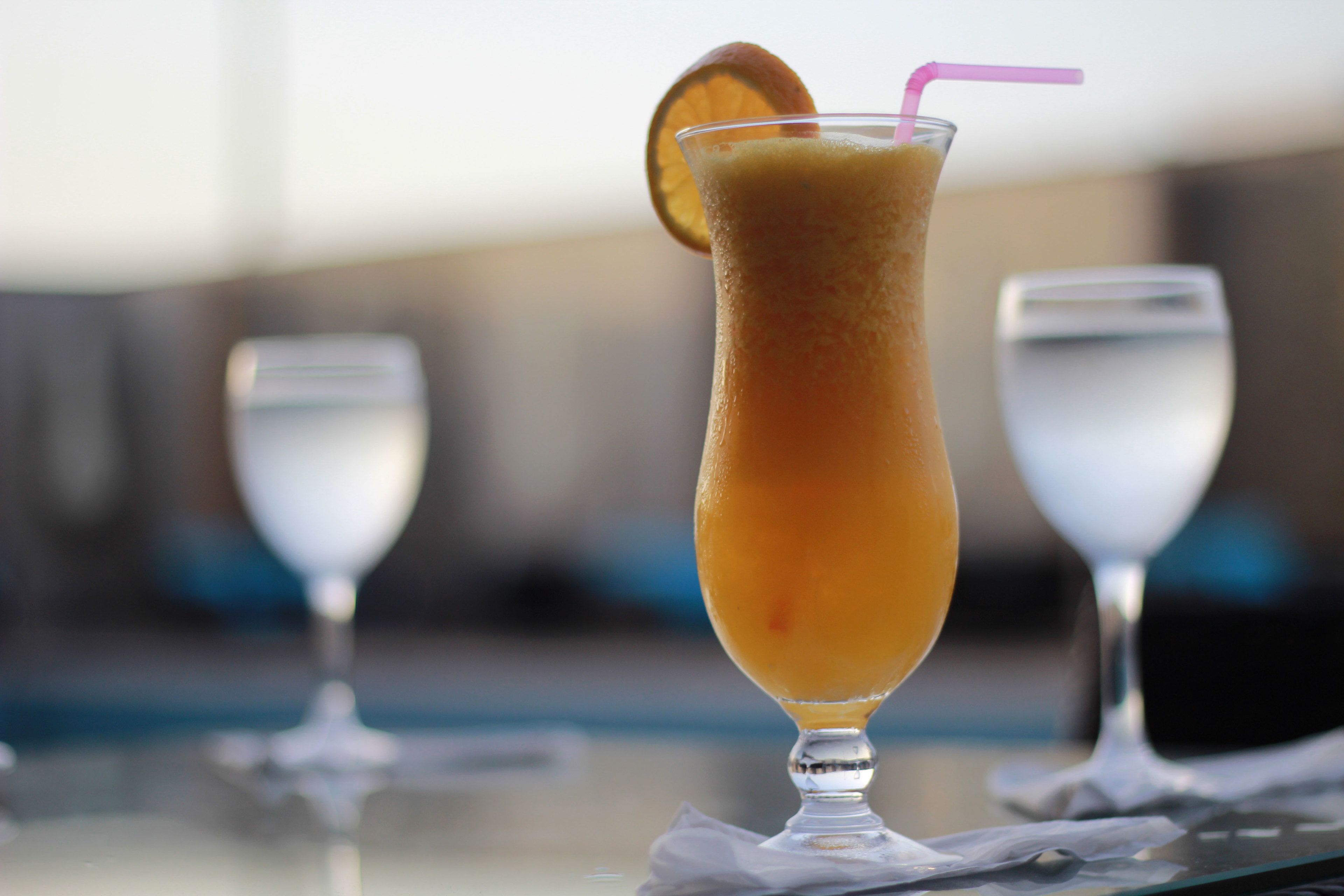 A tall glass with a pink straw and an orange slice garnish, placed on a table with two clear glasses in the background.