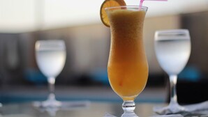 A tall glass with a pink straw and an orange slice garnish, placed on a table with two clear glasses in the background.