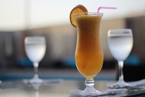 A tall glass with a pink straw and an orange slice garnish, placed on a table with two clear glasses in the background.
