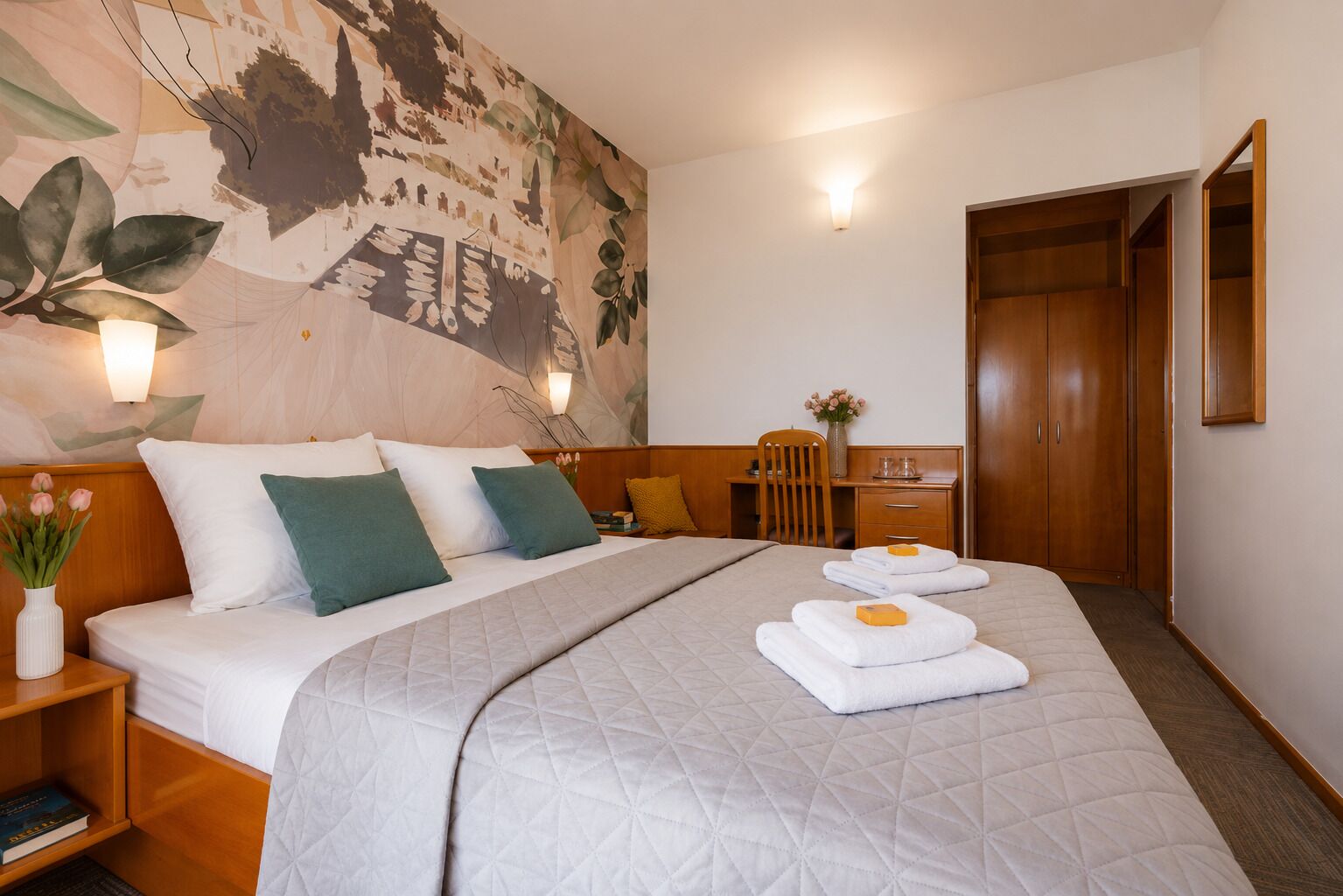 A hotel room with a bed, bedside tables, a desk, a chair, and a wardrobe.