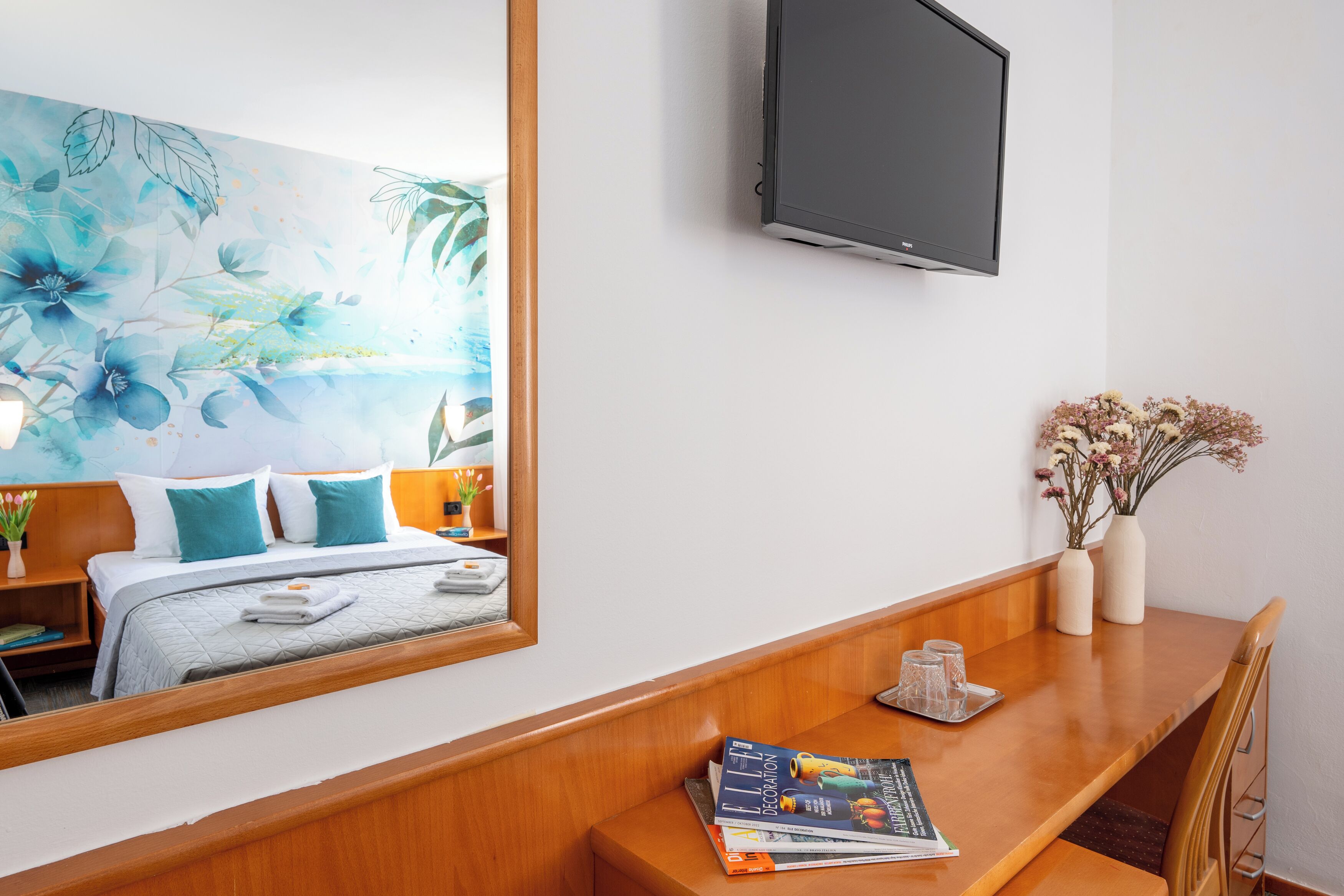 Ein Hotelzimmer mit Bett, einem Holztisch, einem an der Wand montierten Fernseher und einem großen Spiegel.
