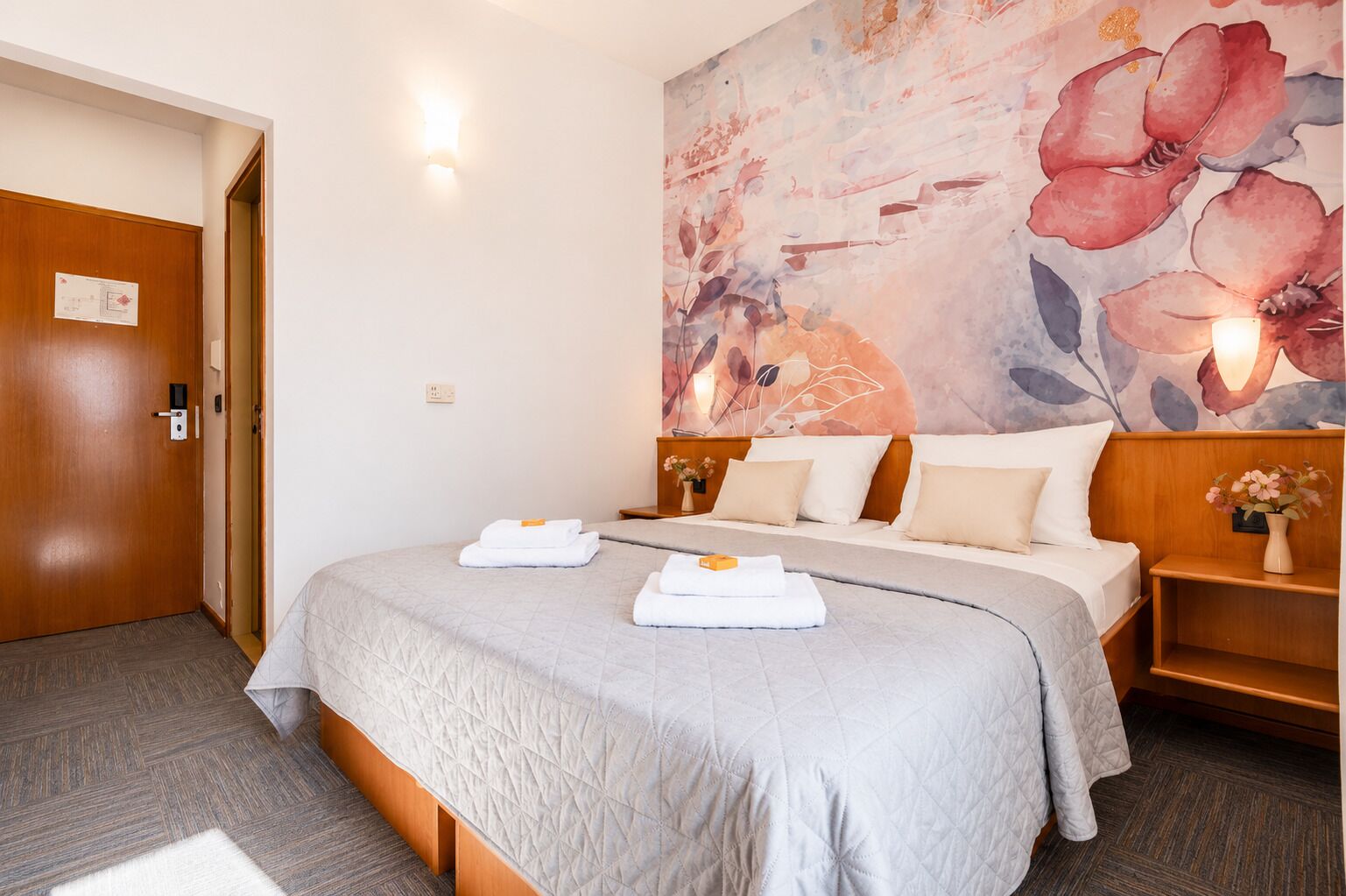 A hotel room with a bed, a nightstand, a wall with a floral painting, and a door.