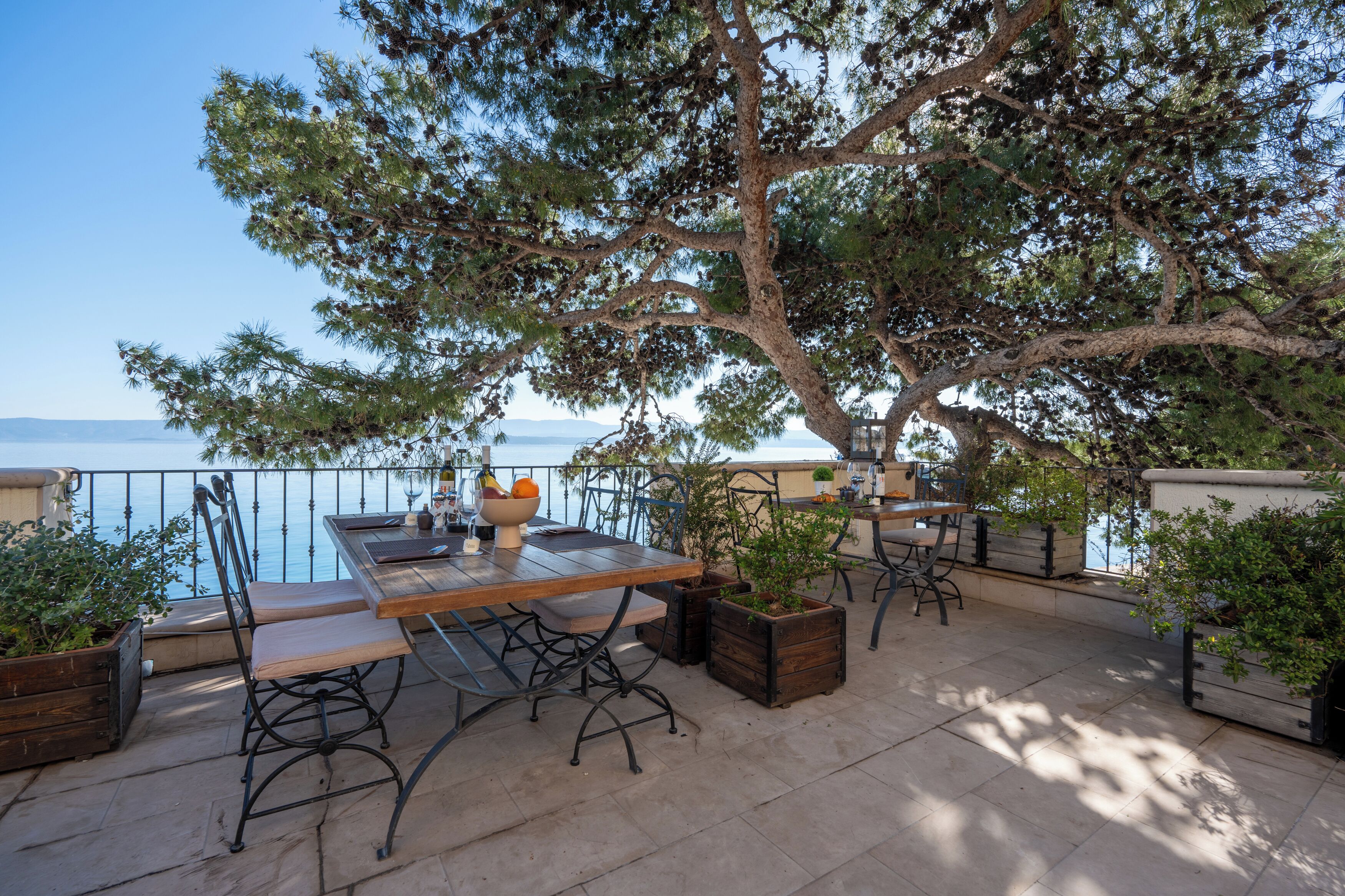 An outdoor patio with a table and chairs, surrounded by potted plants and overlooking a body of water.