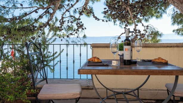 Una mesa con una botella de vino, dos copas y dos cruasanes en una terraza con vistas a un cuerpo de agua.