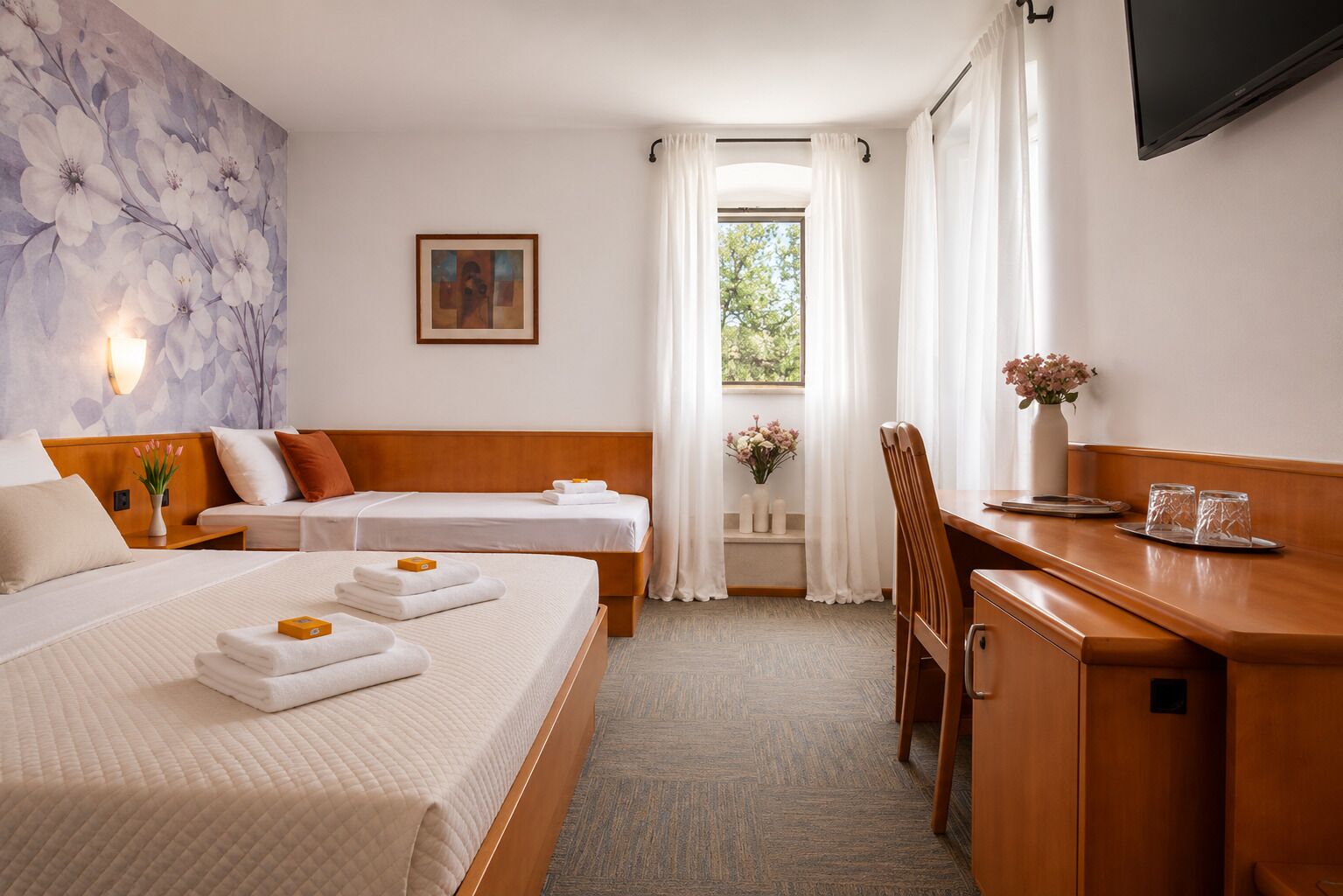 A hotel room with two beds, a desk, a chair, a TV, a window with curtains, a painting on the wall, and a vase with flowers.