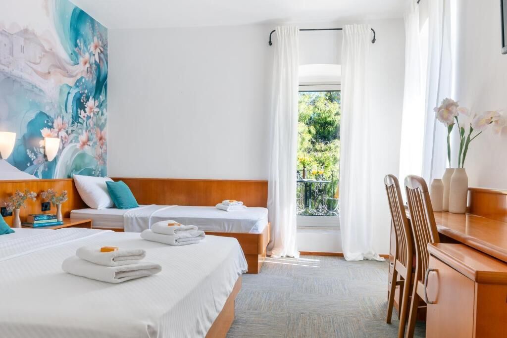 A hotel room with two beds, a desk, a chair, a balcony with a view, and a painting on the wall.