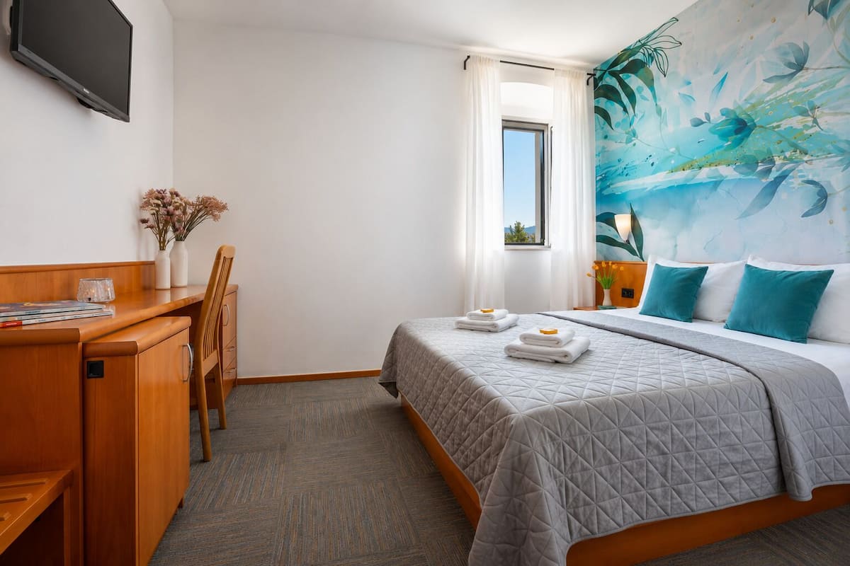 A hotel room with a bed, a desk, a chair, a TV, and a window with curtains.