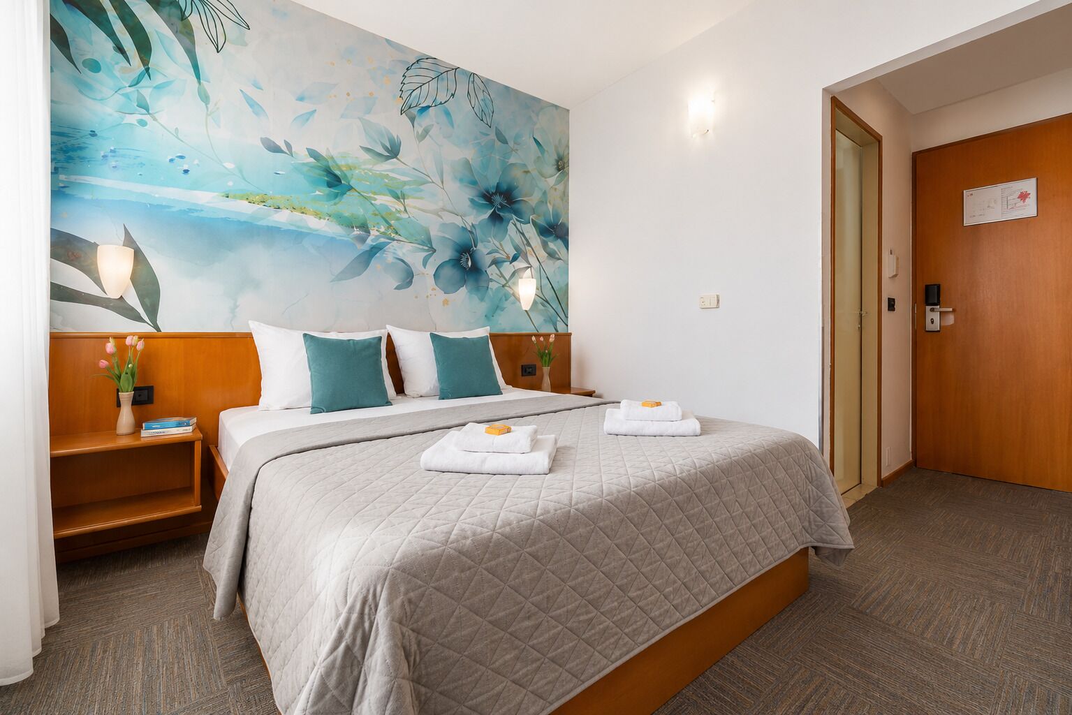 Una habitación de hotel con cama, mesita de noche, un muro con un mural floral, una puerta de madera y una lámpara.