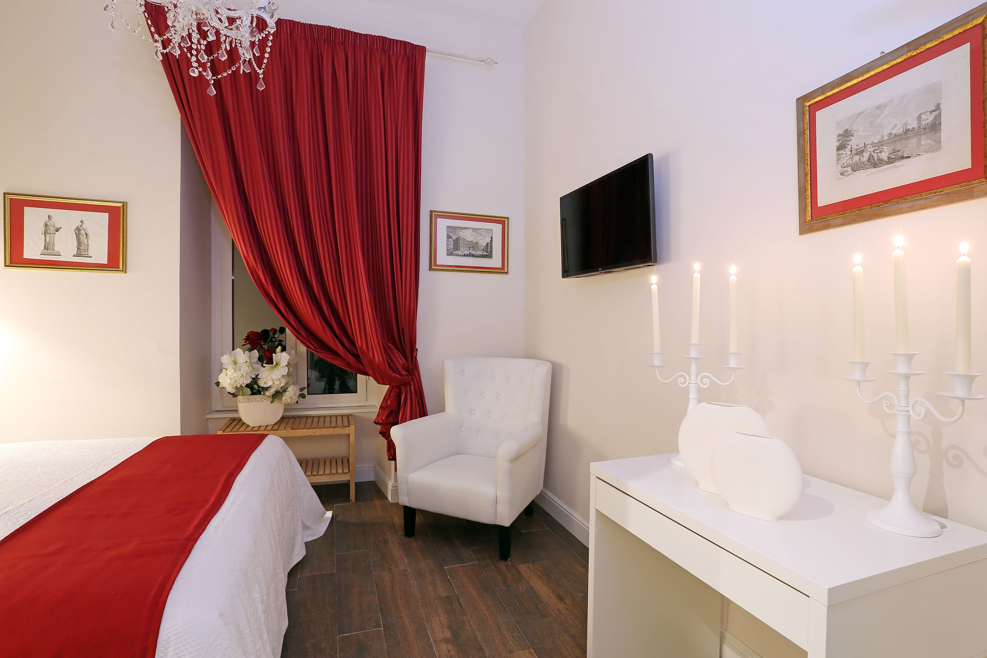 A hotel room with a bed, a white armchair, a TV, framed pictures, and a bedside table with candles.