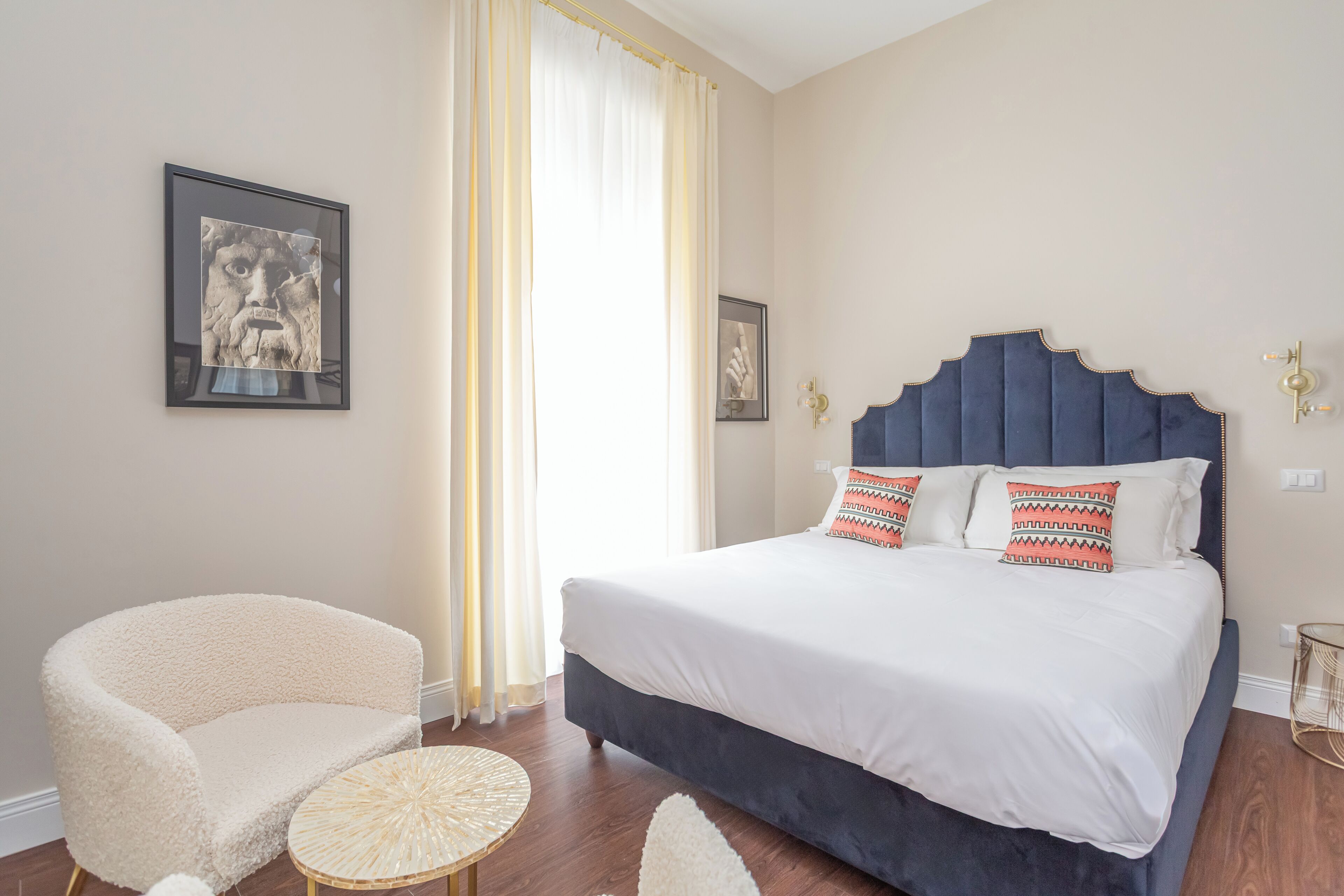 Quarto com cama, uma cadeira, uma mesa redonda e duas fotos emolduradas na parede.