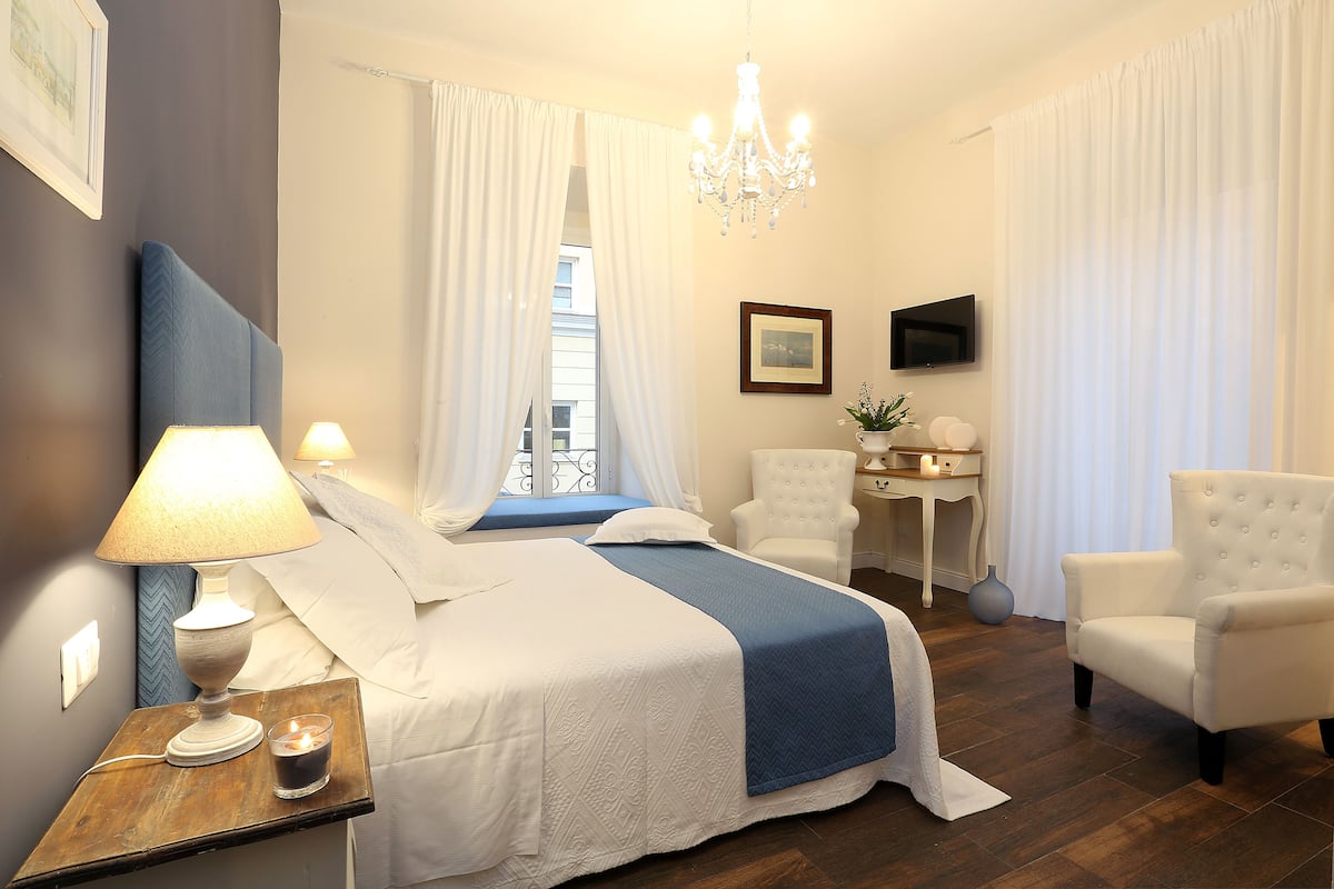 A hotel room with a bed, two chairs, a bedside table with a lamp, a TV, and a window with curtains.