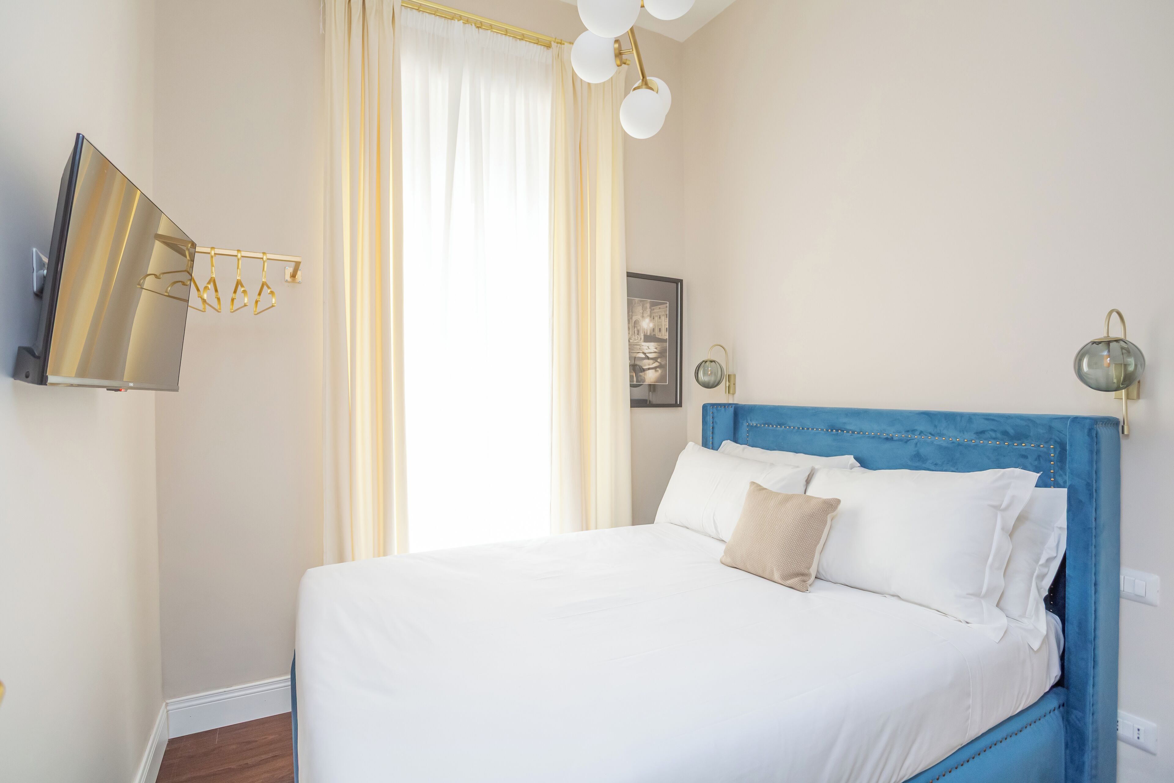 A neatly made bed with white linens and a single beige pillow, a blue headboard, and a wall-mounted TV.