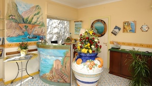 A room with a mural of a coastal scene, a large decorative vase with flowers, a wooden cabinet, and a small table with a vase. - Locanda Del Fiordo (Furore)