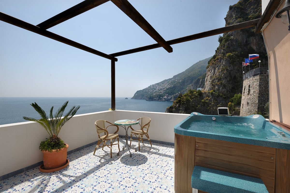 A balcony with a hot tub, potted plant, and wicker chairs overlooking a cliff and the sea.