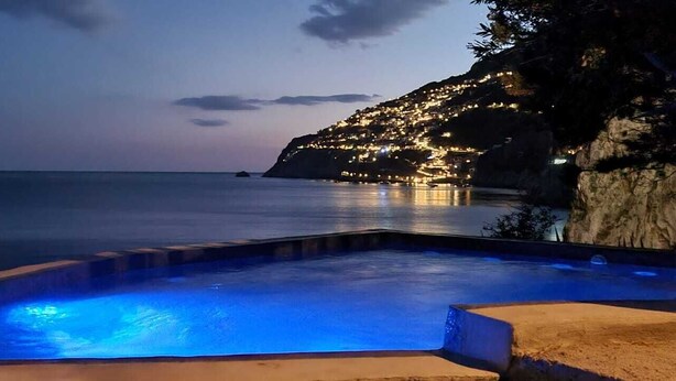 A swimming pool with blue lighting, a coastal view, and a hillside with lights. - Locanda Del Fiordo (Furore)