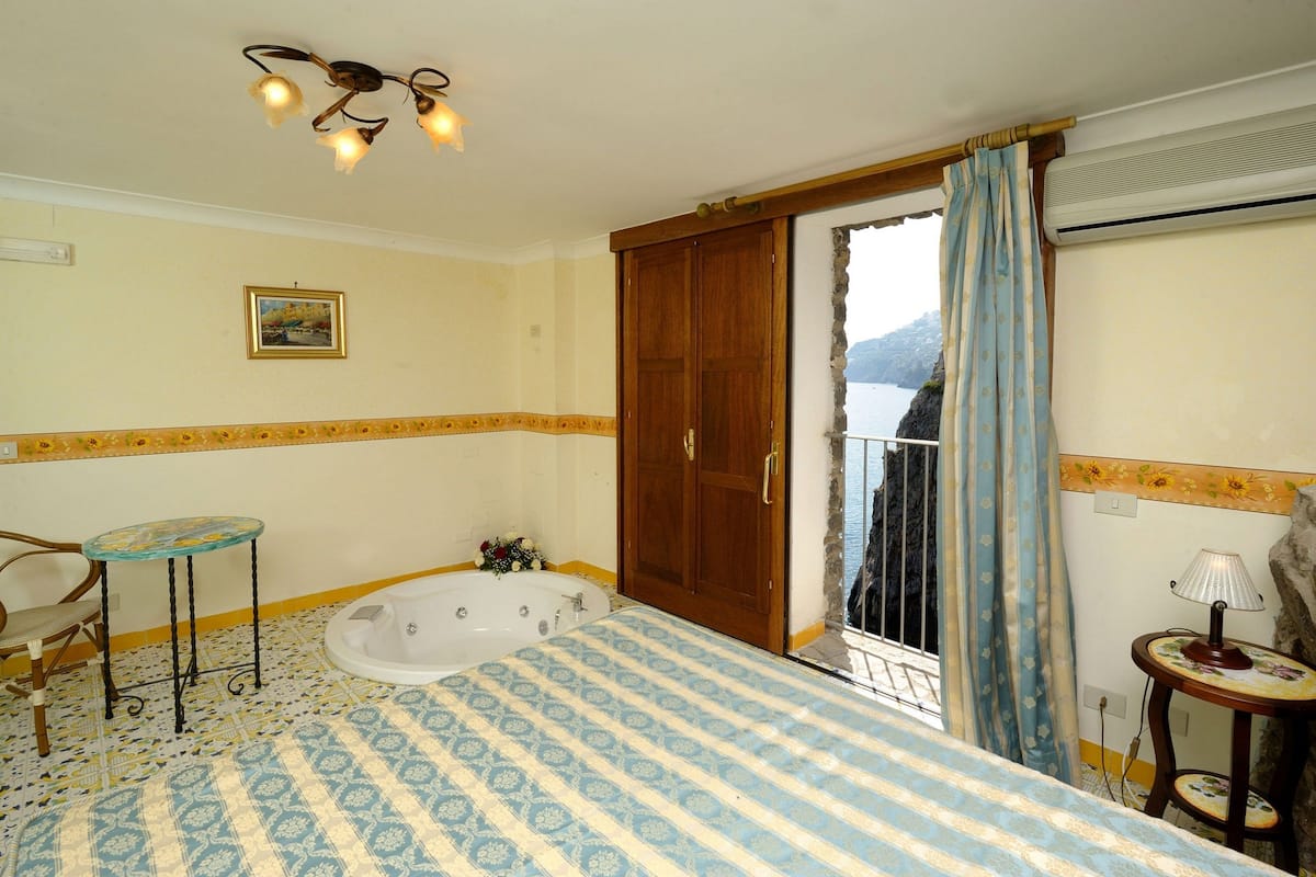 A bedroom with a large bed, a wooden door, a small table with a lamp, and a view of the outdoors.