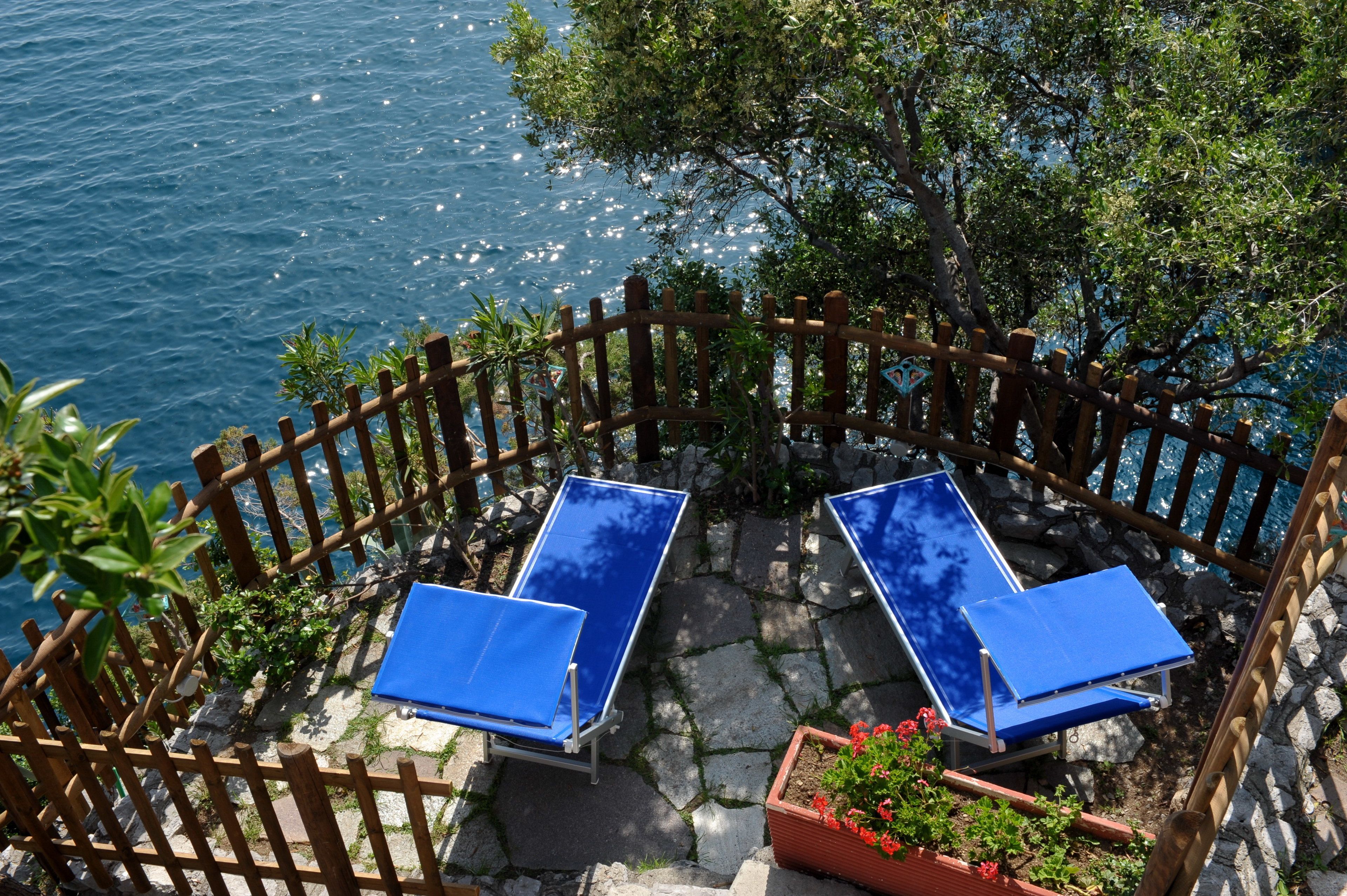 A terrace with two blue sun loungers, a wooden fence, and a view of the sea.