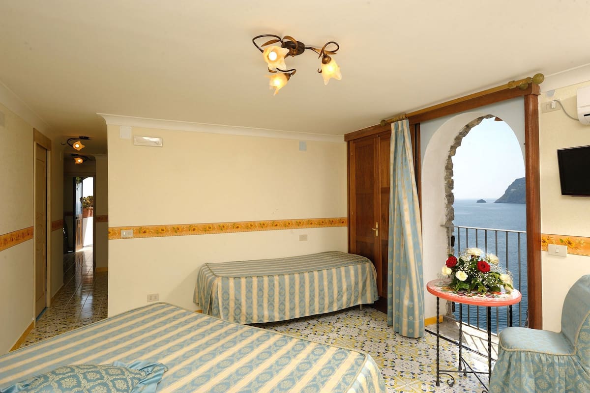 A hotel room with two beds, a small table with a flower arrangement, a balcony with a view of the sea, and a ceiling light fixture.