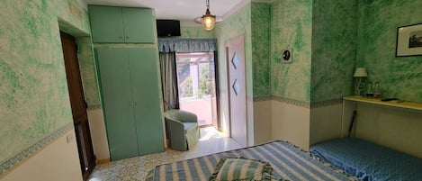 A room with a bed, a wardrobe, a small table with a lamp, and a view of the outdoors.