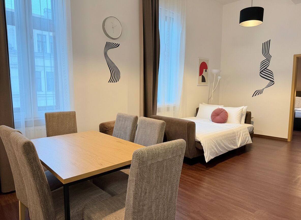 A modern hotel room with a wooden dining table, two sofas, a bed with white bedding, and abstract wall art.