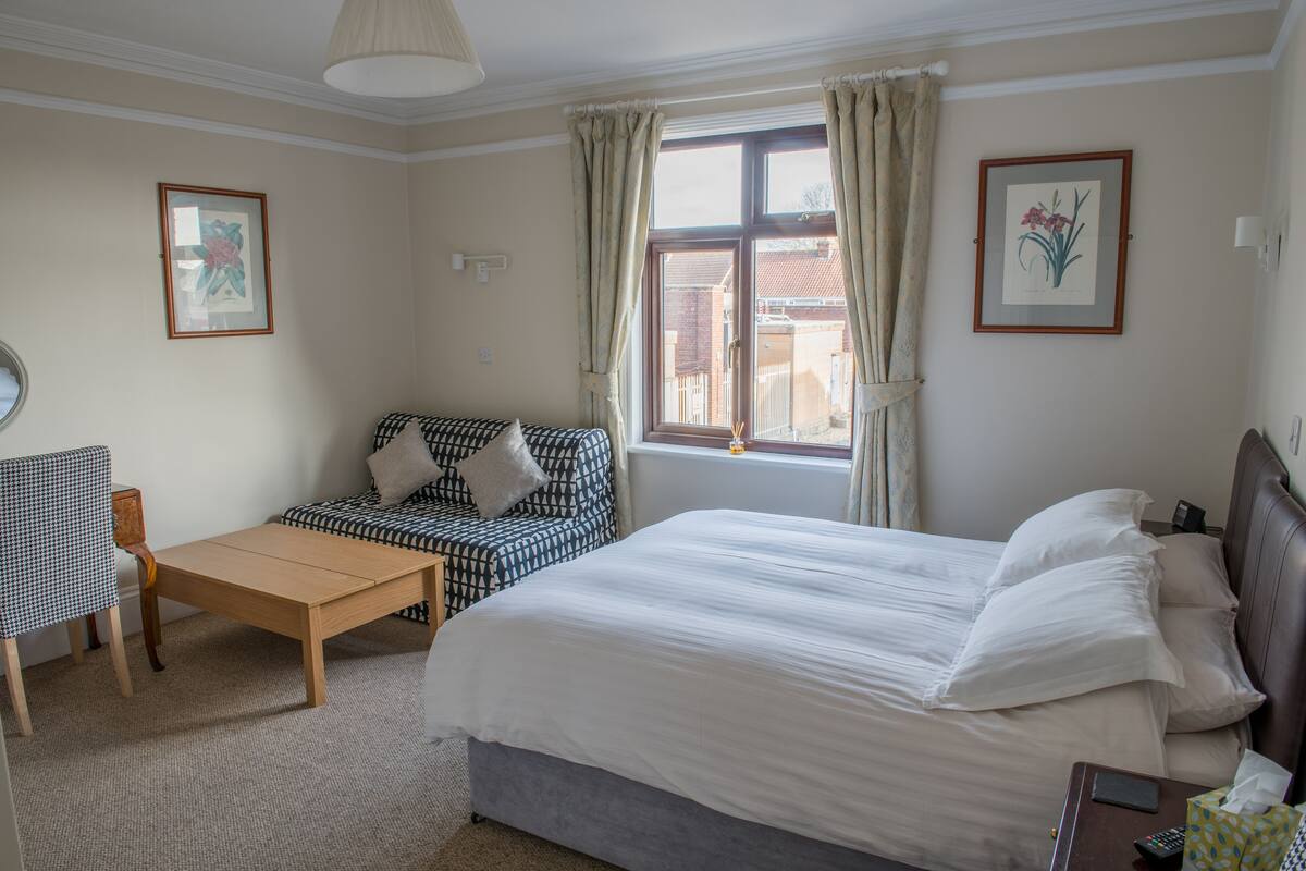A hotel room with a bed, a sofa, a table, and two framed pictures on the walls.