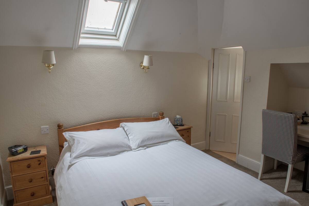 A neatly made bed with white linens, a bedside table with a lamp, a small bedside table with a phone, and a chair with a checkered pattern.