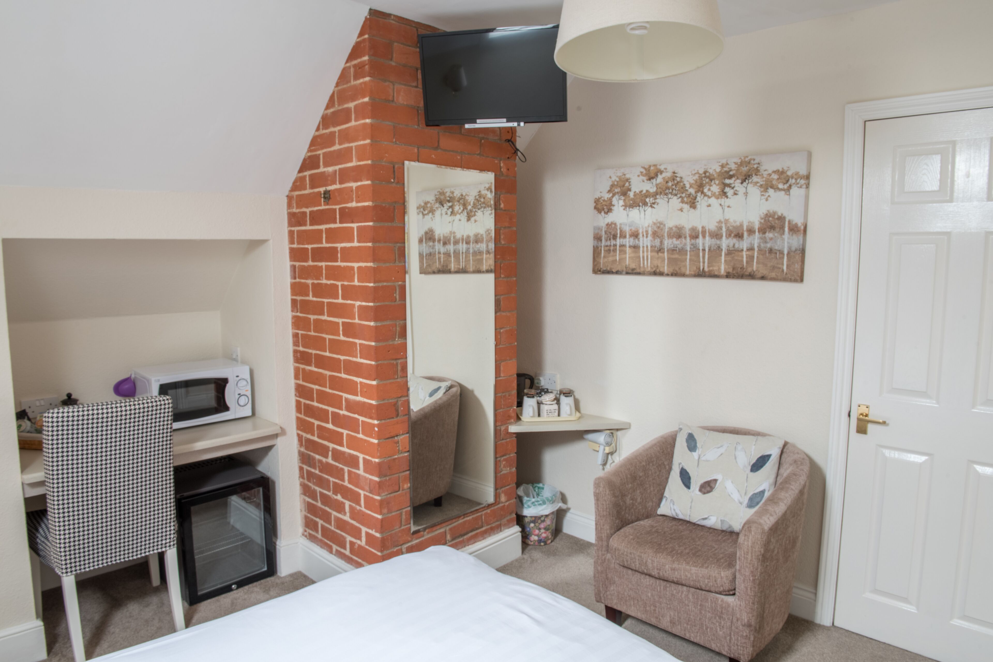 A compact hotel room with a brick fireplace, a television mounted above it, a microwave, a small refrigerator, a chair with a cushion, and a door.