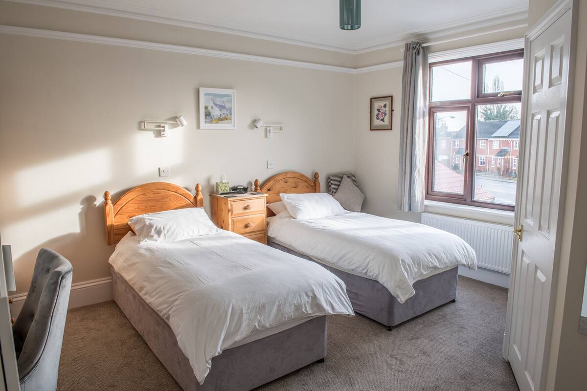 A bedroom with two single beds, a bedside table, a chair, and a window with a view of a street.