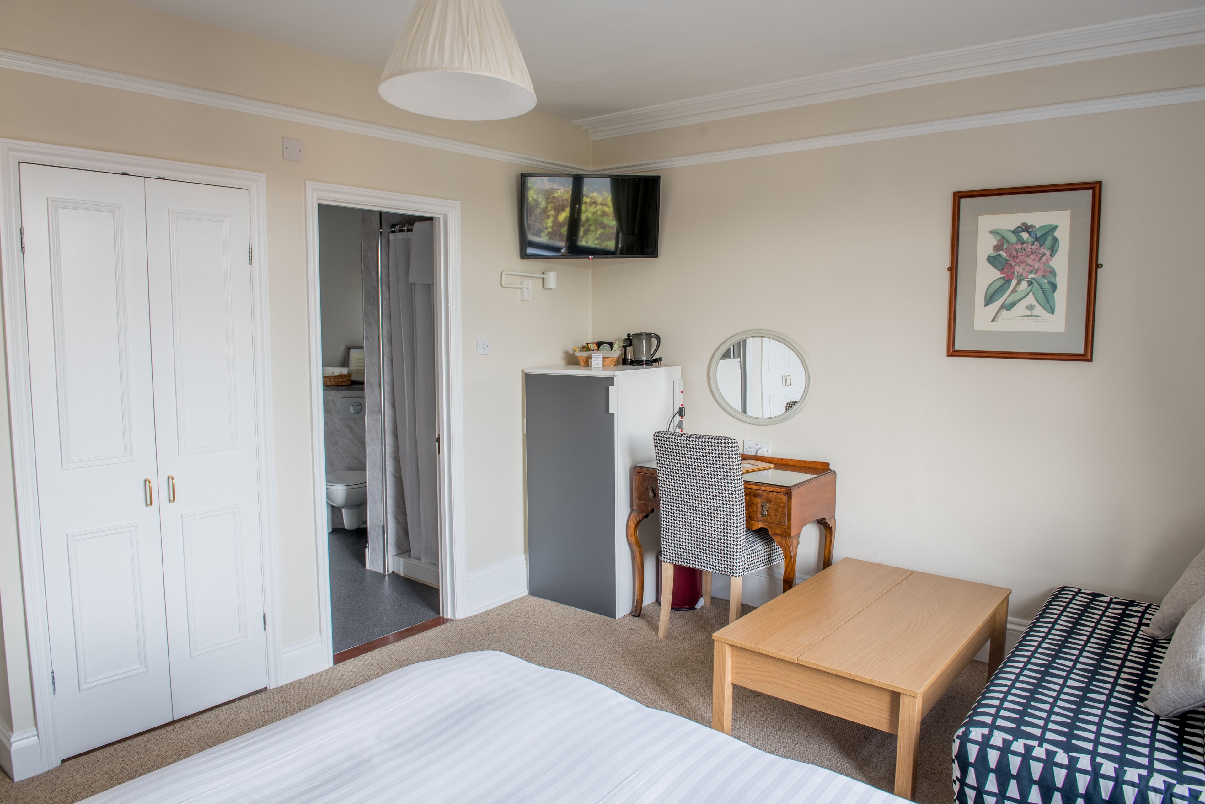 Quarto com cama, escrivaninha, cadeira, mesa, espelho, televisão e um quadro na parede.