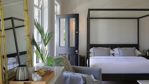 Een slaapkamer met een hemelbed, een nachtkastje met een lamp, een spiegel en een raam met uitzicht op groen.