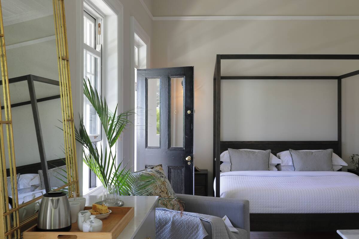 Een slaapkamer met een hemelbed, een nachtkastje met een lamp, een spiegel en een raam met uitzicht op groen.