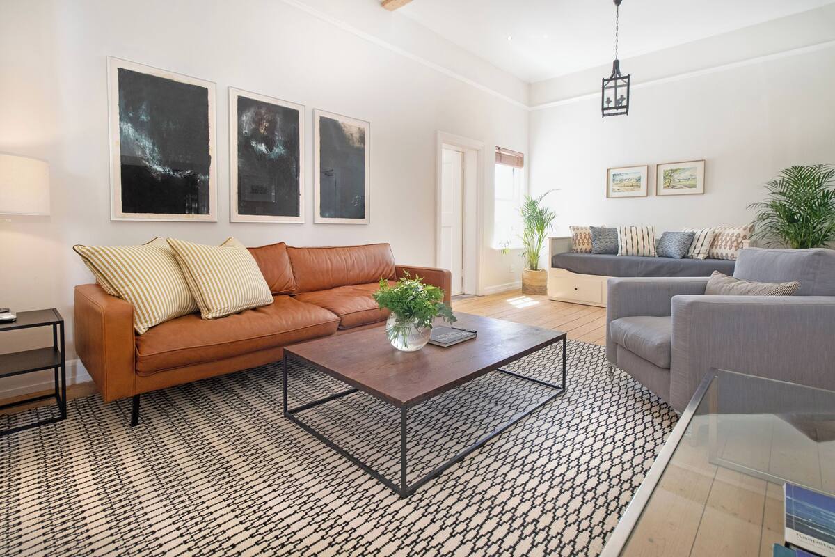 Een moderne woonkamer met een bruine leren bank, een grijze bank, een gekleurd vloerkleed en kunstwerken in lijsten aan de wanden.