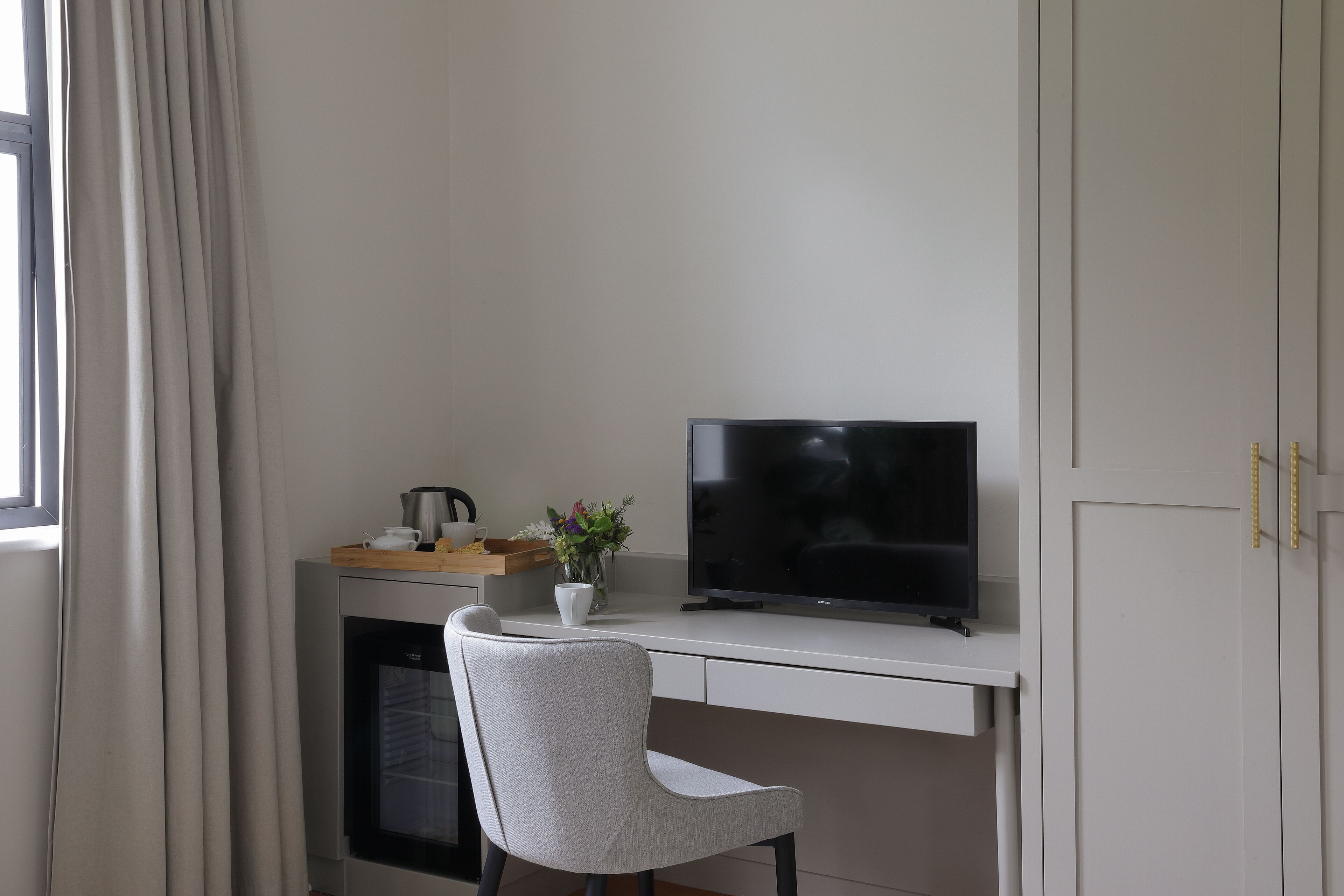 Una habitación moderna con un escritorio blanco, un televisor de pantalla plana, una silla gris, una planta pequeña y una cafetera.