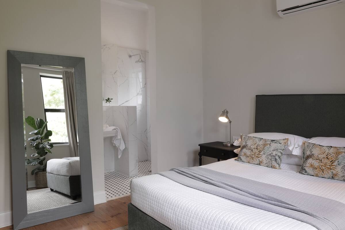 Een slaapkamer met een bed, nachtkastje, spiegel en een badkamer door een geopende deur zichtbaar.