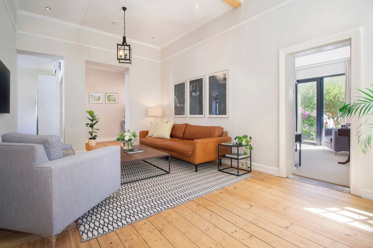 Un salón amplio con un sofá gris, un sofá de cuero marrón y una mesa de centro. Hay tres cuadros enmarcados en la pared, una lámpara y una planta en maceta.
