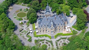 Eine Vogelperspektive eines großen, historischen Gebäudes mit einer symmetrischen Fassade, umgeben von üppigem Grün und einem kreisförmigen Teich.