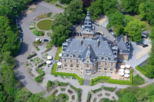 Vista aérea de un edificio grande e histórico con una torre destacada, rodeado de jardines cuidados y un área de estacionamiento.