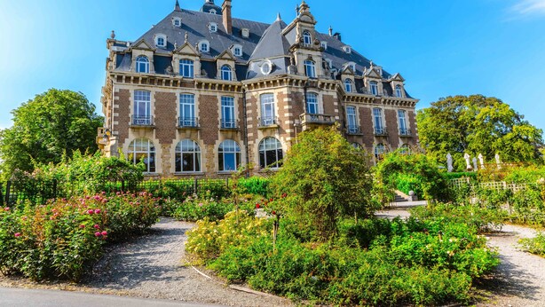 Un edificio grande e histórico con múltiples ventanas y fachada simétrica, rodeado de jardines y senderos bien cuidados.