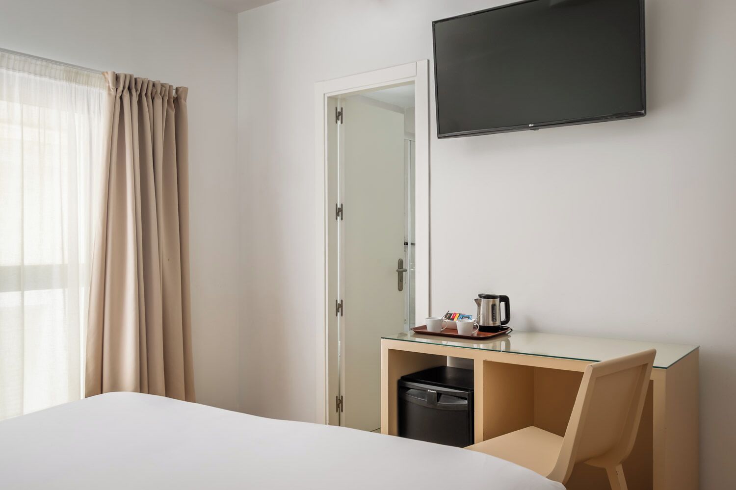 A hotel room with a bed, a desk with a coffee maker, a trash bin, a television, and a window with curtains.
